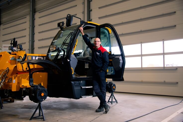 Man poseert bij gele heftruck in werkplaats.